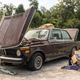 a maroon 1974 bmw 2002tii sits on a concrete pad in a field with its hood and trunk open. the car has been abandoned for decades; it's covered in dirt and grime and surrounded by tools, jacks, and tow straps, all of which were used to pull it out of the field where it sat for 25 years